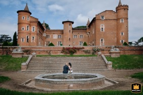 In front of the Château de Pibrac, Haute-Garonne, the newlywed couple sits on the edge of a fountain, in a charming scene with the majestic château as their backdrop.